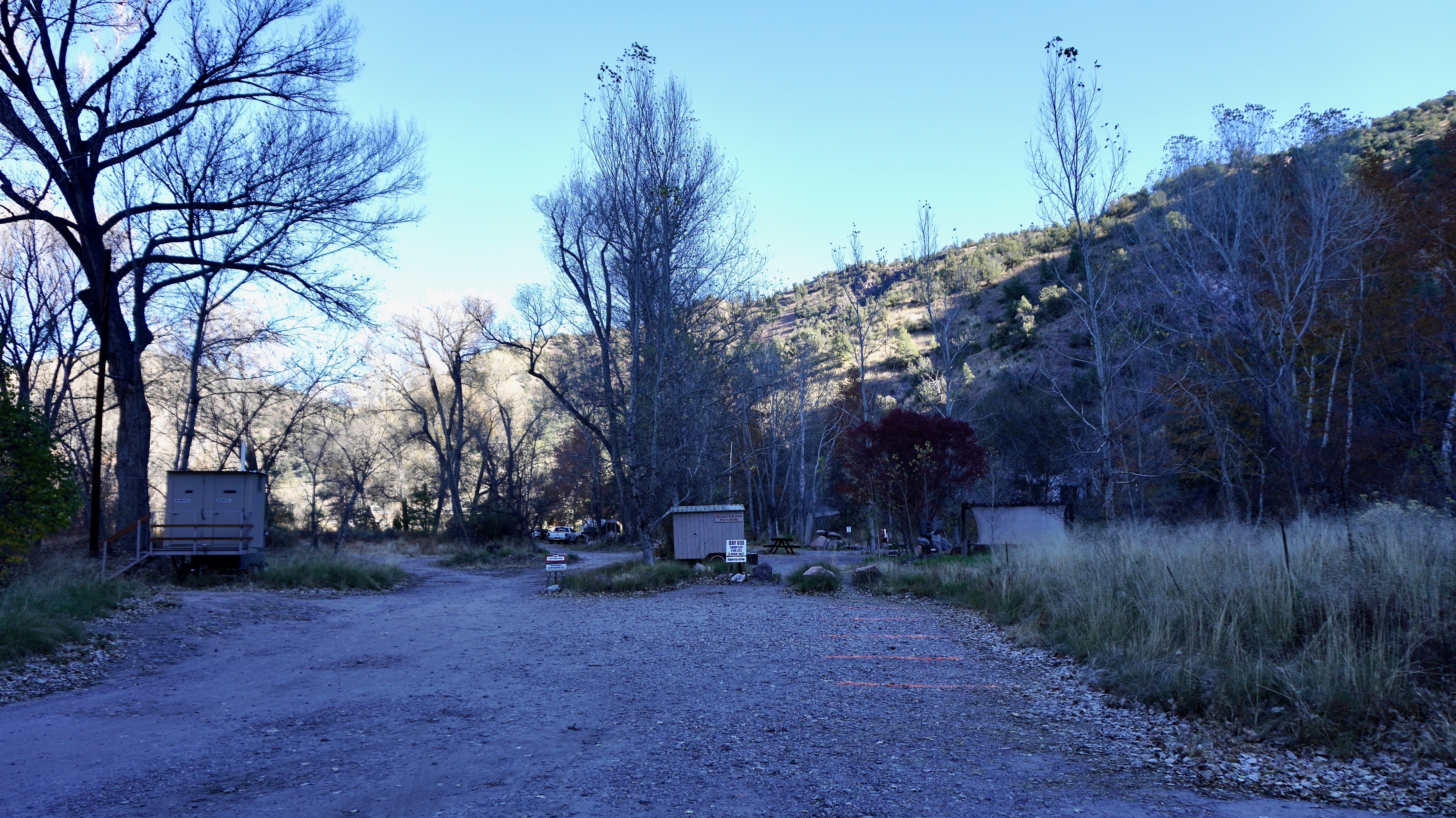 a large gravel area for parking with the toilet building to the left, check in booth at center, pools to the right with grass trees and cliff around