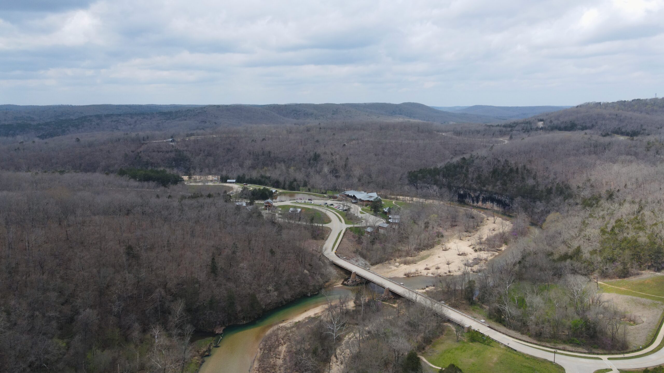 a drone picture over looking the echo bluff state park lodge, a road, bridge and green creek cutting through a forested area with hills