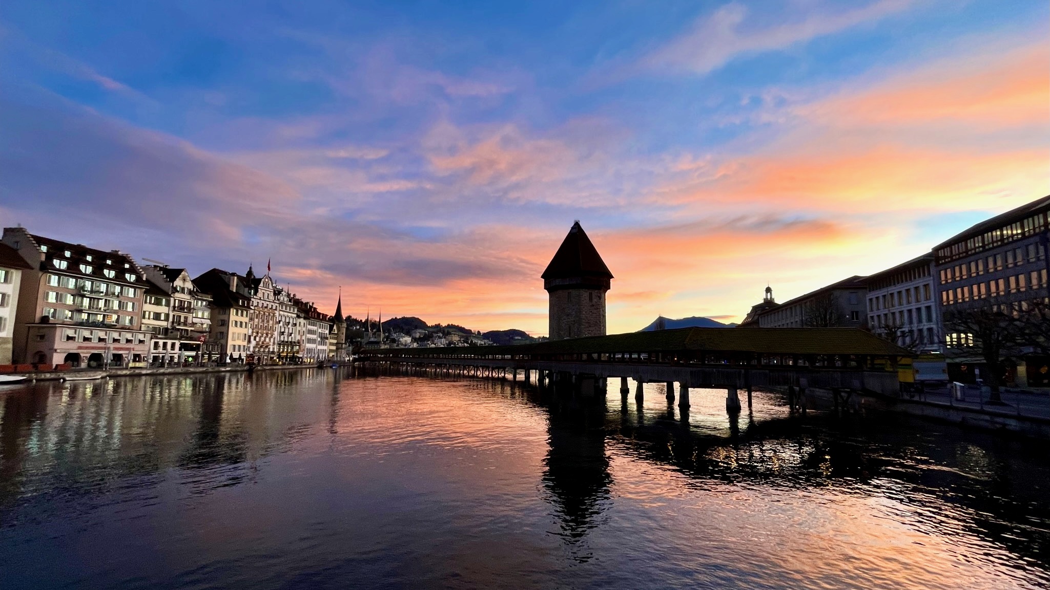 a blue and pink sky with the chapel bridge in the foreground with buildings on both sides and the river below in lucerne