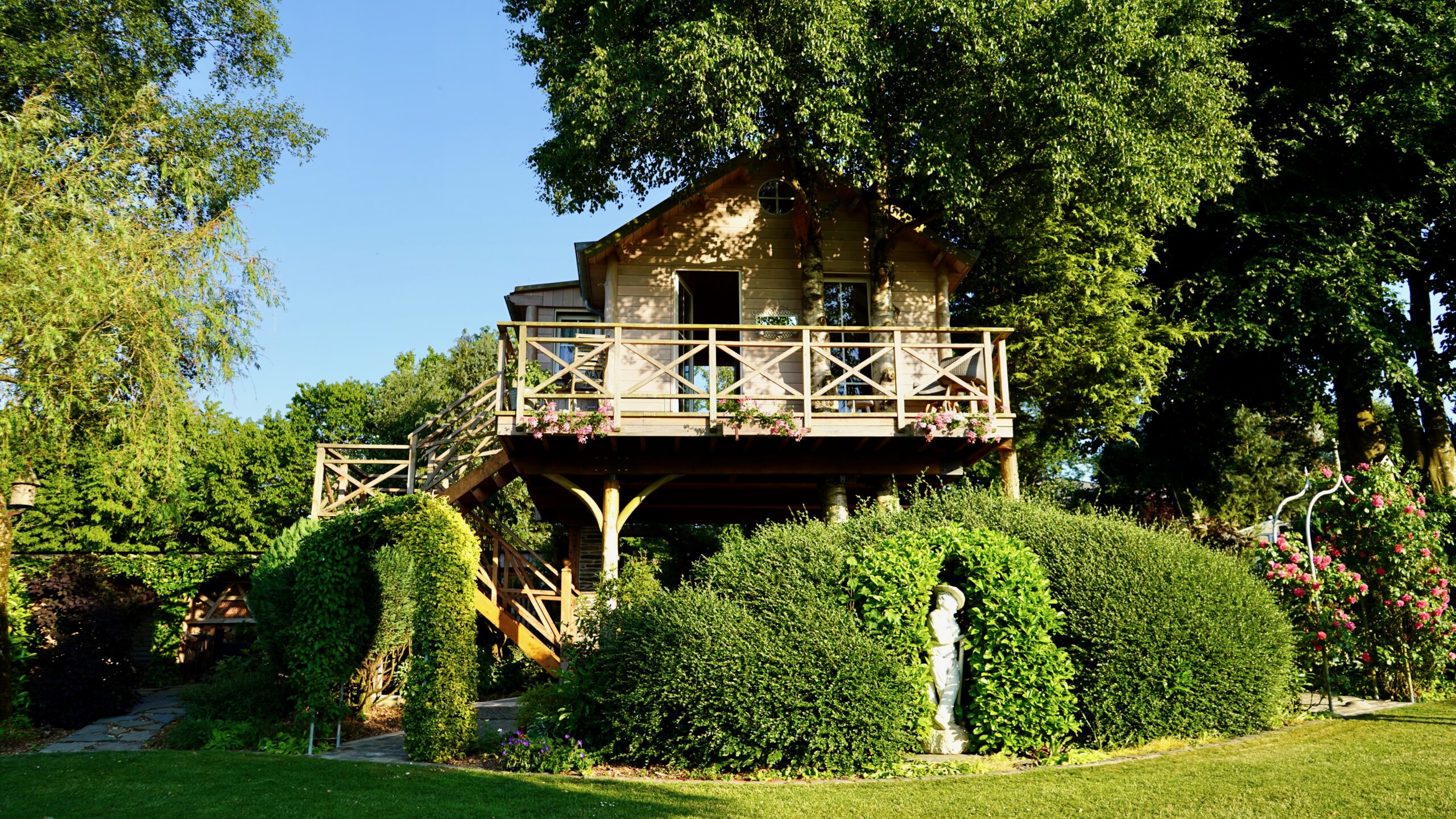 a small wooden tree house airbnb with bushes and statues in front on the ground with a railing and flowers all around