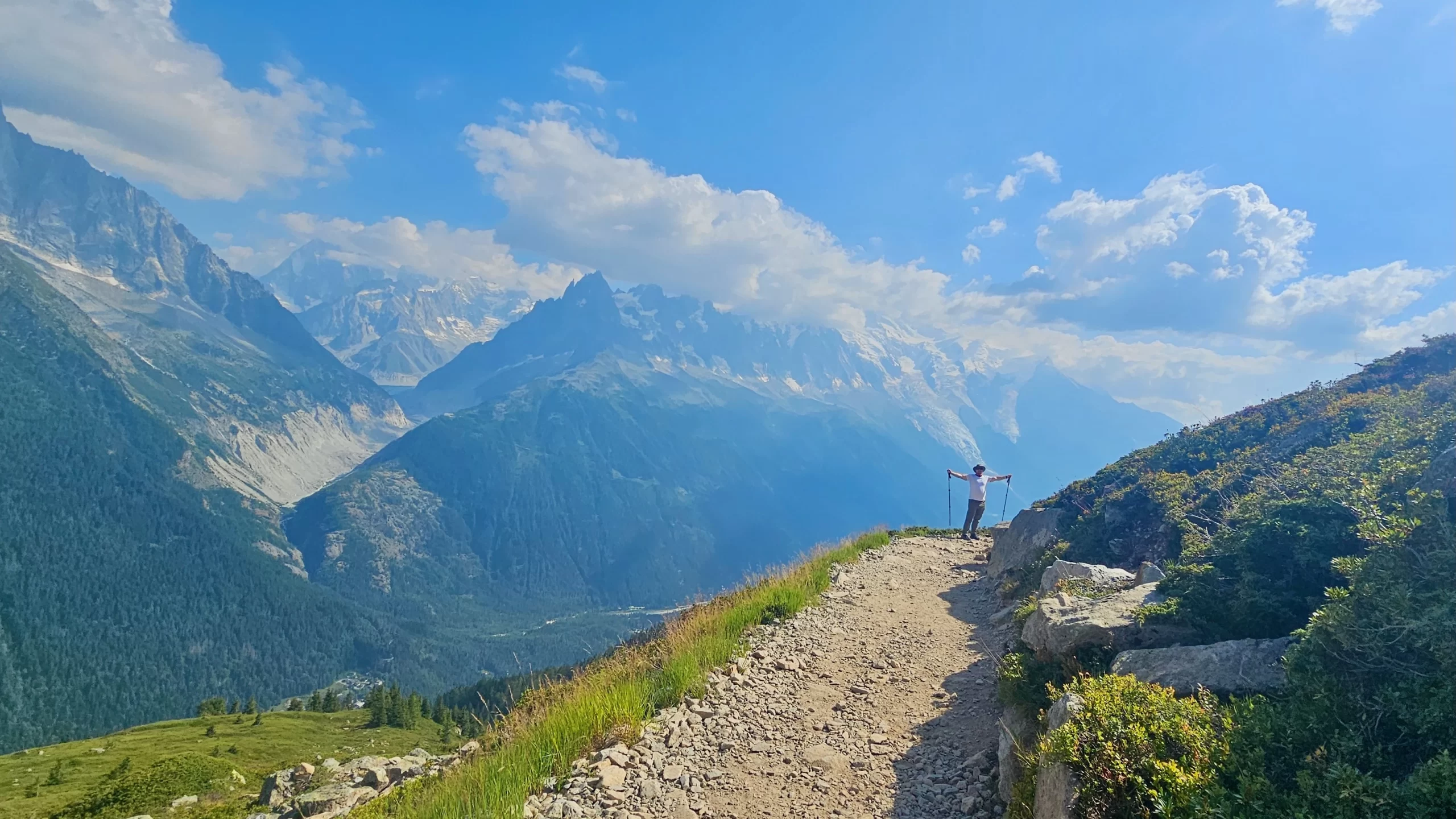 a man in white shirt arms wide standing on the tour du Mont Blanc trail with mountains and cloud behind