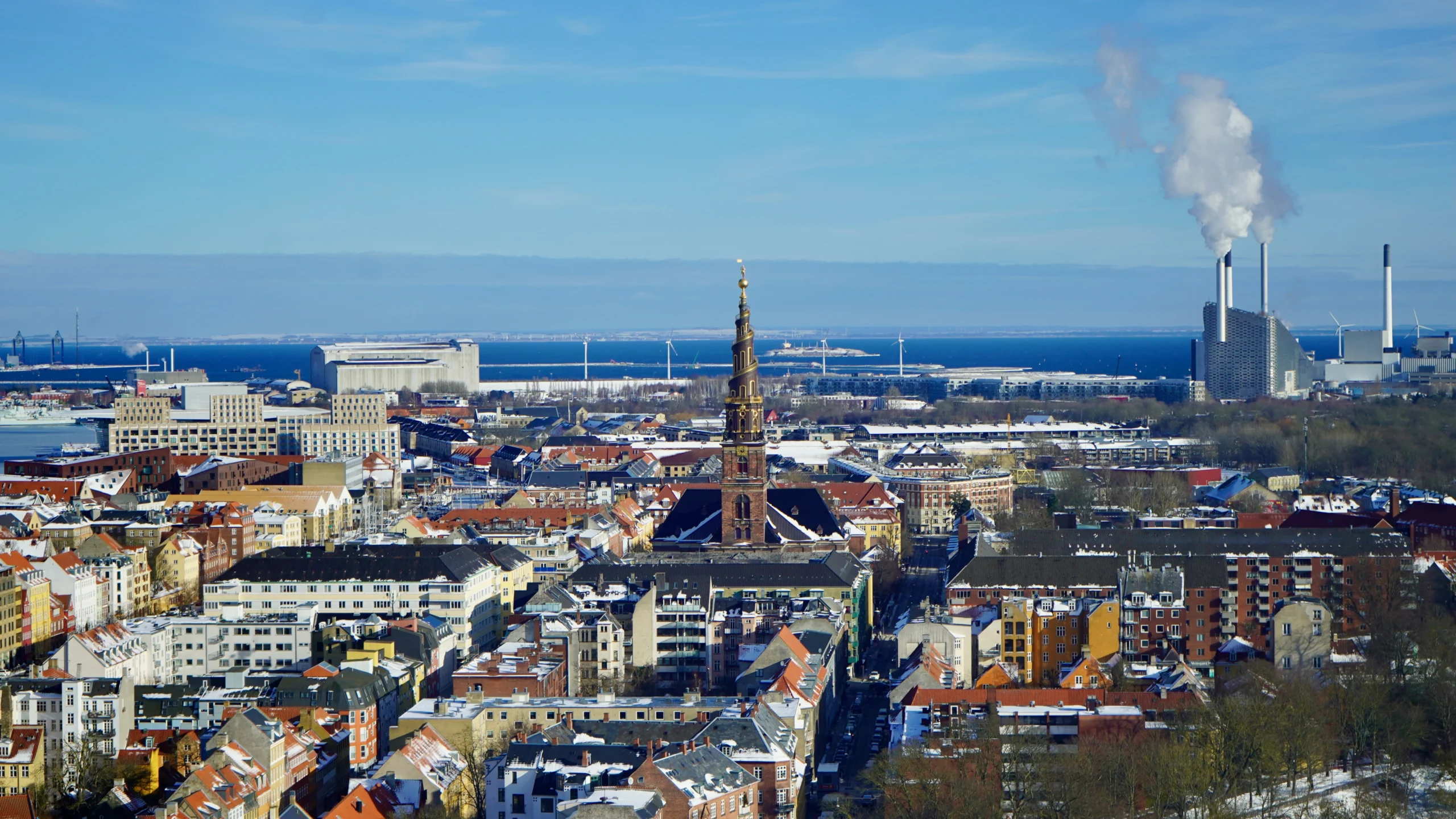 a high vantage point photo from the front of the Church of Our Saviour in copenhagen taken from far away with city all around, power plant in the back right, and blue sunny sky.